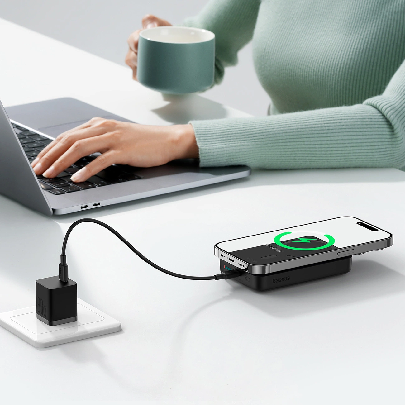 Powerbank Baseus Magnetic Mini w biurowej scenerii leży obok laptopa na biurku i jest podłączony do kontaktu przez kabel a na nim znajduje się telefon który jest ładowany