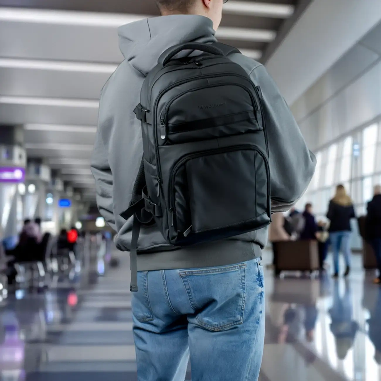 A young man with a Wozinsky WPW-20L backpack on his back stands at the airport, with a crowd visible in the background.