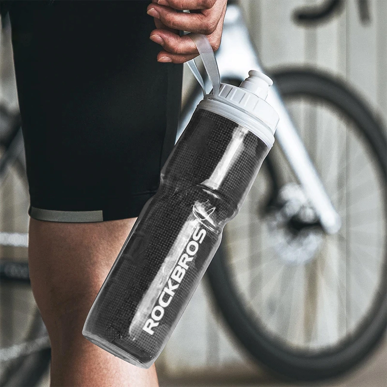 A man holding the handle of a Rockbros 35210019001 bicycle water bottle along his body. There is a bicycle in the background