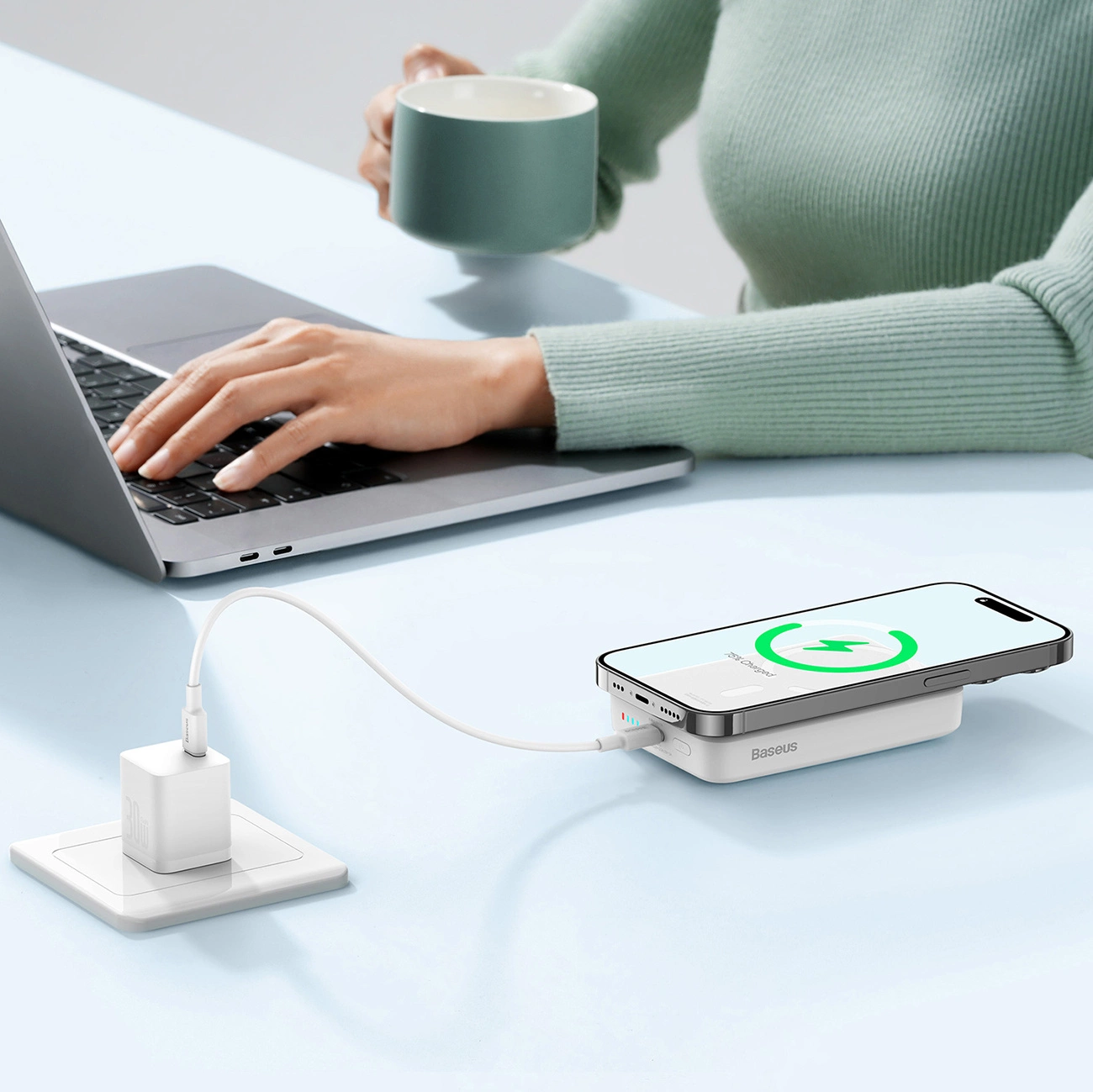 The Baseus Magnetic Mini powerbank in an office setting lies next to the laptop on the desk and is connected to the socket via a cable, and there is a phone on it that is being charged.