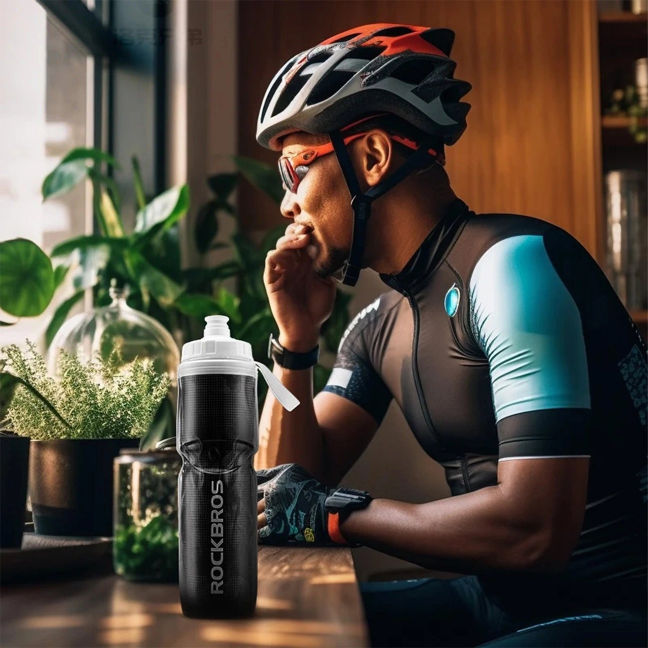 A man in cycling clothes and a helmet sitting by the window, on the table nearby there is a Rockbros bicycle bottle 35210019001